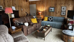 A cozy living room with a brown sofa, a blue couch, and a grey armchair. There's a coffee table with a laptop, a lamp, and a small table with a bowl and remote controls. - Round Lake's Happy Swimmers 1970's-80's Retro Cottage (Waupaca)