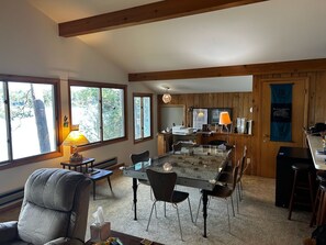A spacious living area with a glass-top dining table, a comfortable sofa, and a kitchen area with wooden cabinets and a large window. - Round Lake's Happy Swimmers 1970's-80's Retro Cottage (Waupaca)