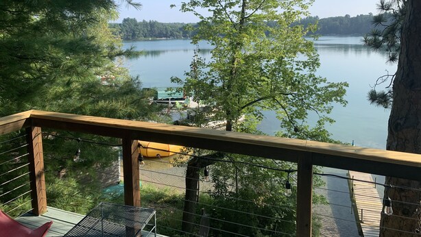 A wooden deck with a view of a lake, surrounded by trees. - Round Lake's Happy Swimmers 1970's-80's Retro Cottage (Waupaca)