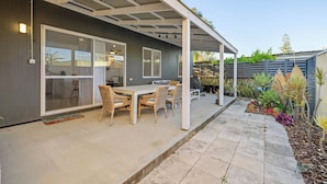A covered patio with a dining table and chairs, a sliding glass door leading to a kitchen, and a garden with various plants and a surfboard. - Sou Wester - is a bright, light and modern holiday home. (Lancelin)