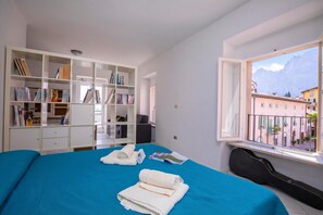 A bedroom with a bed, a bookshelf, a window with a view of buildings, and a guitar. - Dogana Best Lake View, Torbole sul Garda, Italy (Torbole sul Garda)