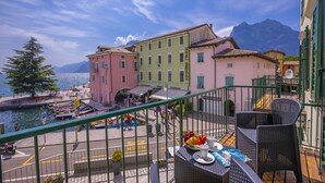 A balcony with wicker furniture, a table with a fruit bowl and cups, and a view of colorful buildings and a mountain in the background. - Dogana Best Lake View with Terrace, Torbole sul Garda, Italy (Torbole sul Garda)