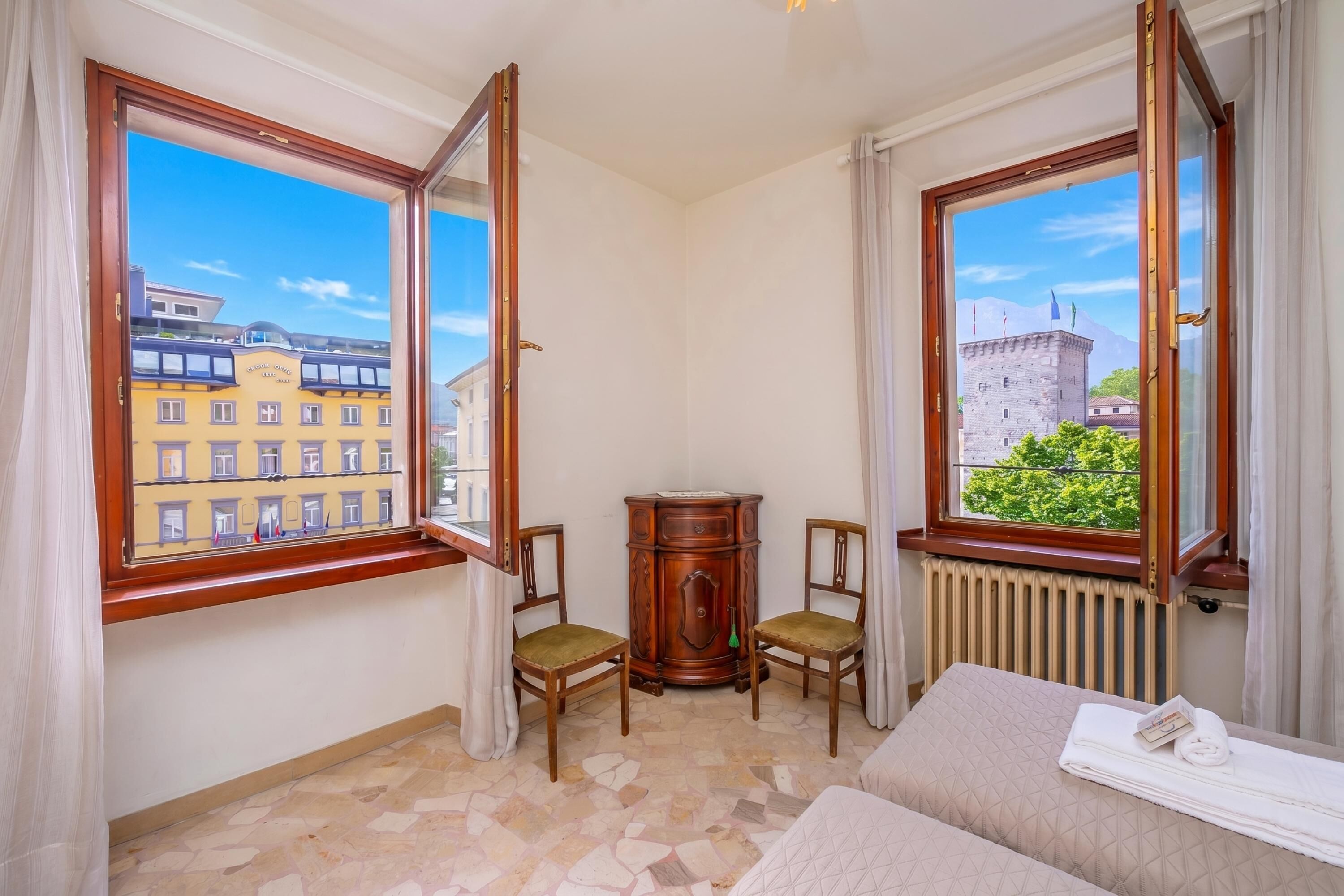 Camera d'albergo con un'ampia finestra, un termosifone, un armadietto in legno, due sedie e un letto.