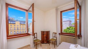 A hotel room with a large window, a radiator, a wooden cabinet, two chairs, and a bed.
