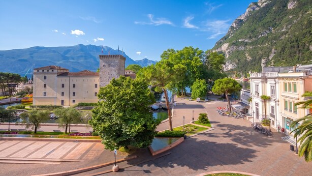 A square with a fountain, a castle-like building, and a mountain in the background.