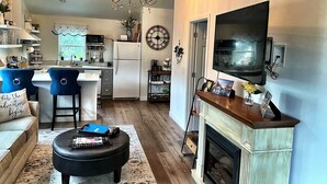 Cocina moderna con isla central, zona de comedor con sillas azules, chimenea y televisor montado en la pared.