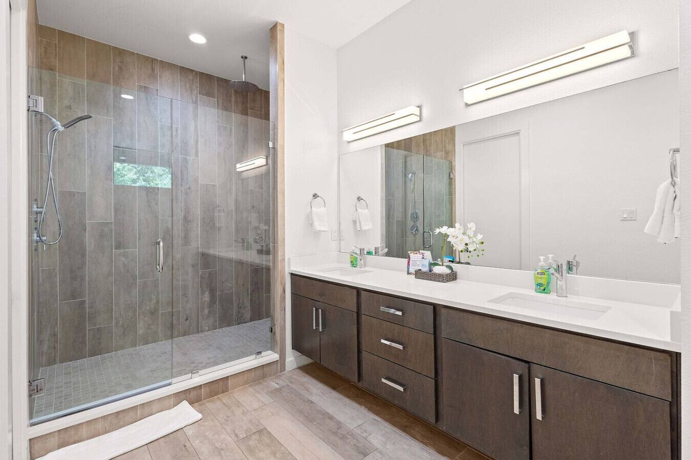 Een moderne badkamer met een douche met glazen wand, een grote spiegel en een wastafelmeubel met donkere kastjes.