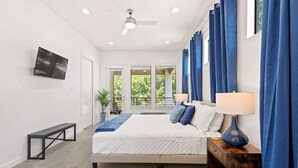 A bedroom with a bed, bedside tables, a bench, a TV, and a view of greenery outside.