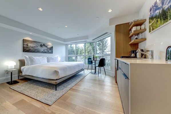 A modern bedroom with a large bed, bedside tables, a desk, and a view of the outdoors through large windows.
