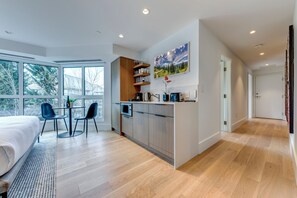 Une chambre moderne avec un grand lit, un coin repas avec des chaises, une cuisine avec un plan de travail et une vue sur les arbres à travers de grandes fenêtres.