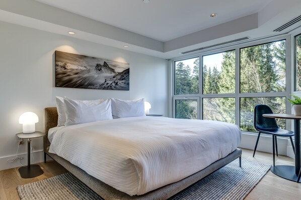 A modern bedroom with a large bed, a painting, and a view of trees through the window.