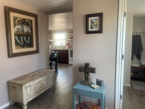 Um corredor com um banco de madeira, um quadro emoldurado, uma pequena mesa com uma cruz e uma porta que dá para um quarto.