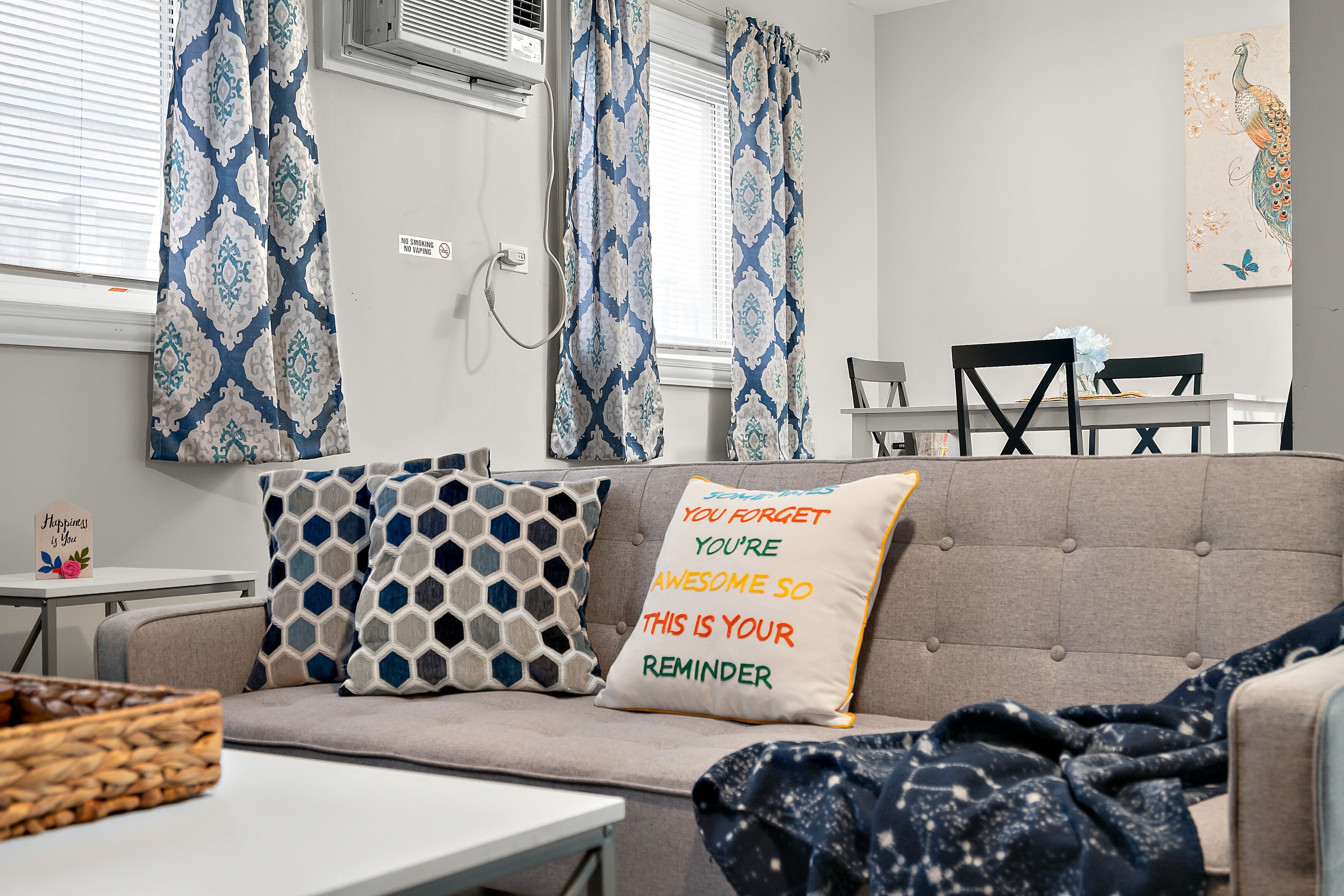 A living room with a grey sofa, patterned cushions, a wall-mounted air conditioner, and a dining area with a table and chairs.