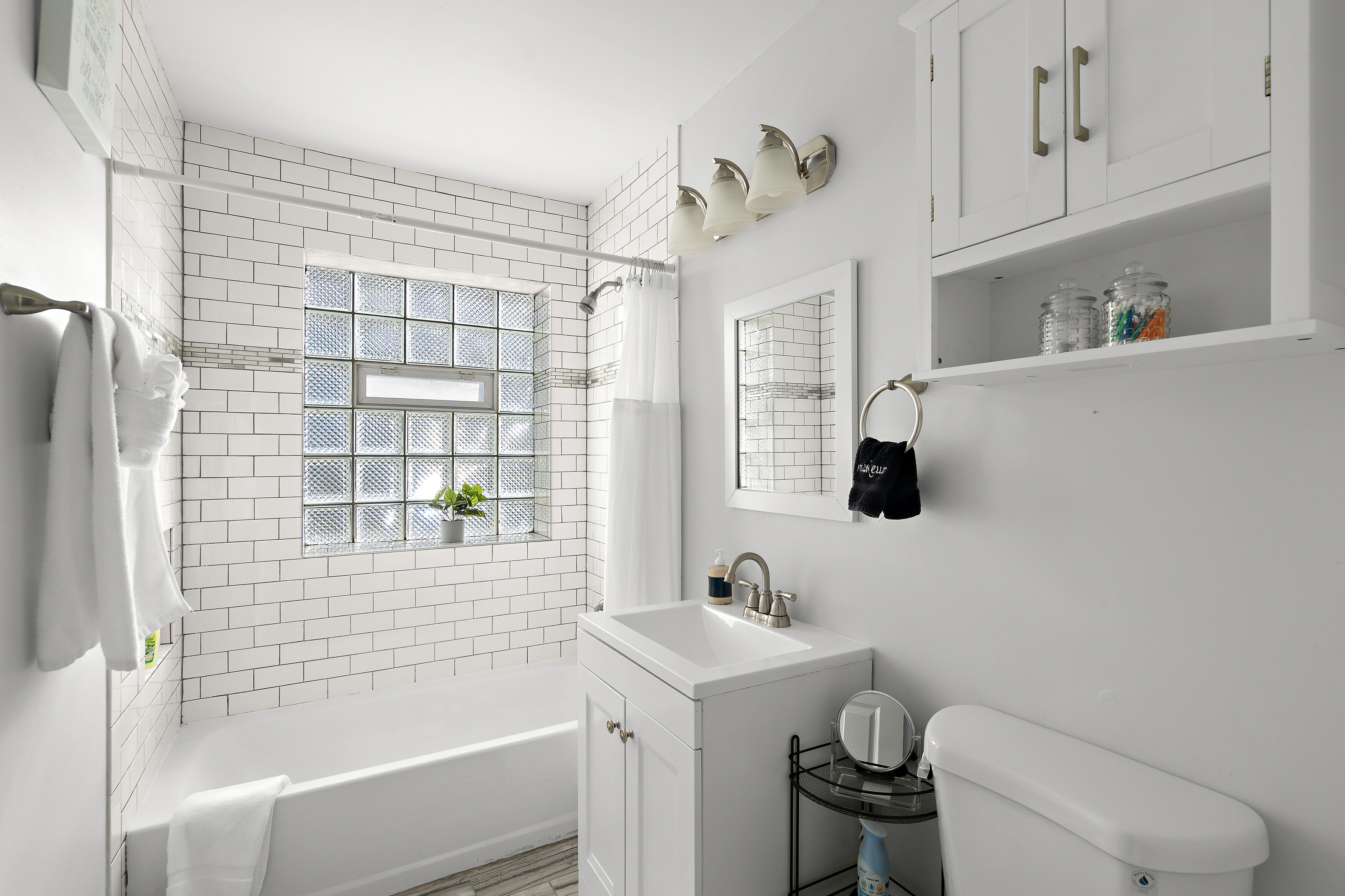 Ein modernes Badezimmer mit weißen Fliesen, einer Badewanne, einem Waschbecken mit Spiegel und einem Fenster mit Aussicht.