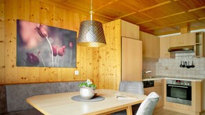 Un intérieur en bois comprenant une table à manger, des bancs, un luminaire suspendu et un coin cuisine avec des placards et des appareils électroménagers.