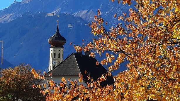 A church with a bell tower, surrounded by autumn foliage and mountains in the background.