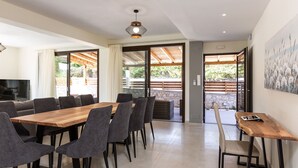Uma sala de jantar com mesa de madeira e cadeiras estofadas em cinza, uma televisão e vista para o exterior através de portas de vidro.