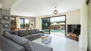 Uma sala de estar moderna com um sofá modular cinzento, uma mesa de centro redonda, uma TV de tela plana e uma grande porta de vidro deslizante que dá para uma área de piscina.