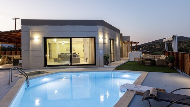 Uma área moderna junto à piscina, com piscina cristalina, zona de estar coberta e um edifício com grandes portas de vidro.