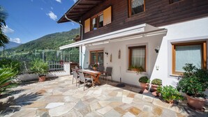 A two-story wooden house with a stone patio, outdoor seating, and potted plants. - Apartment "Sonnenblume" with Mountain View, Pool, Wi-Fi, Balcony & Garden (Rifiano)