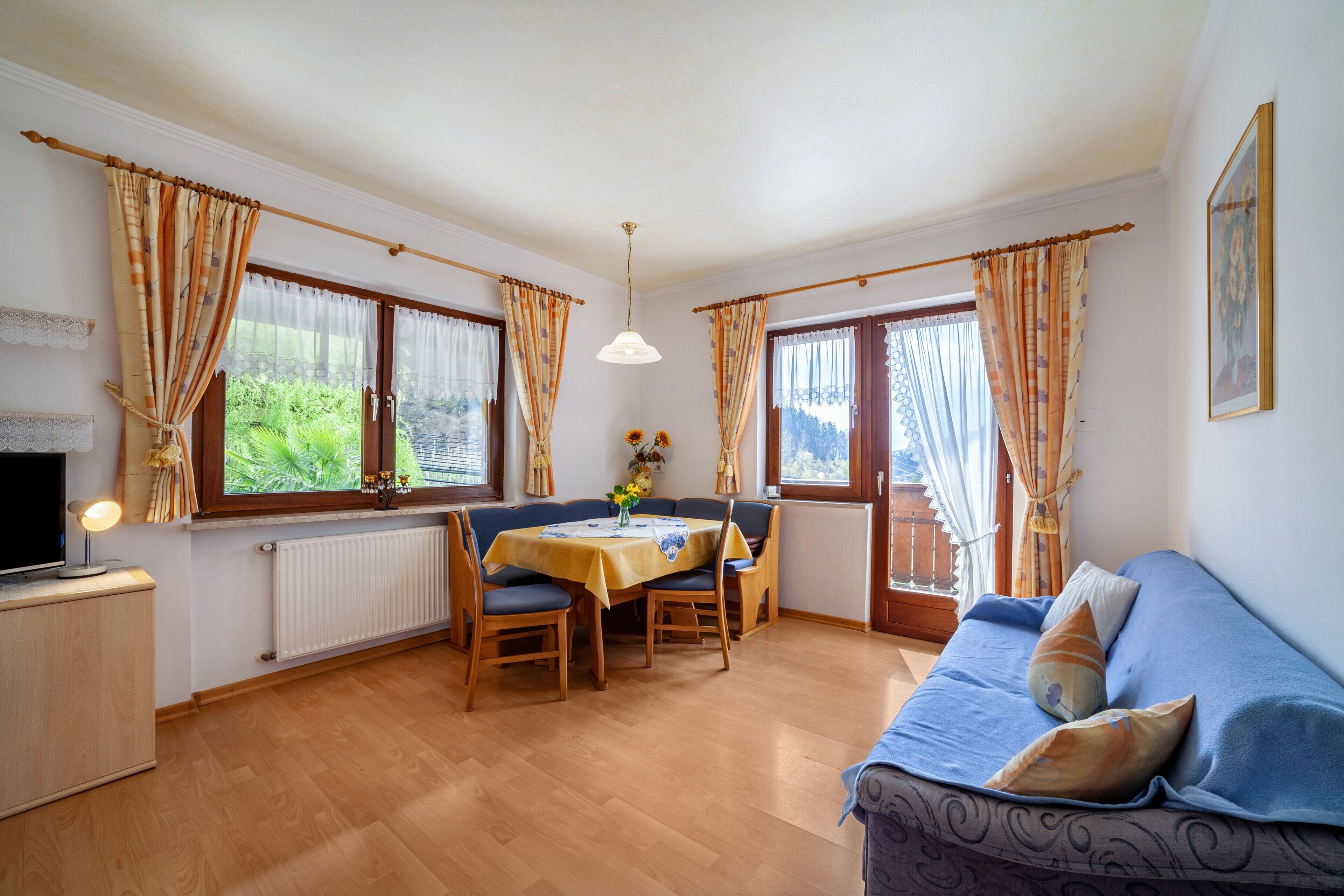 A room with a dining table, chairs, a sofa, a TV, and windows with curtains.