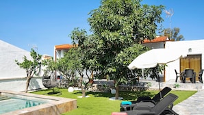 Un área junto a la piscina con sillones de descanso, una piscina pequeña y un jardín con árboles y un edificio blanco al fondo.