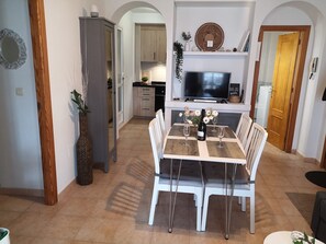 A dining area with a table set for two, a television above the table, and a kitchen in the background.