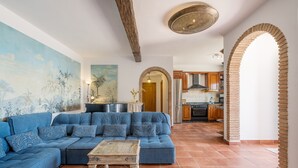 A living room with a blue sofa, a wooden coffee table, a mural, and a kitchen visible through an archway. - Cortijo La Alberquilla – Andalusian Charm with Mountain and Sea Views, Pool, Wi-Fi, and Garden (OTIVAR)