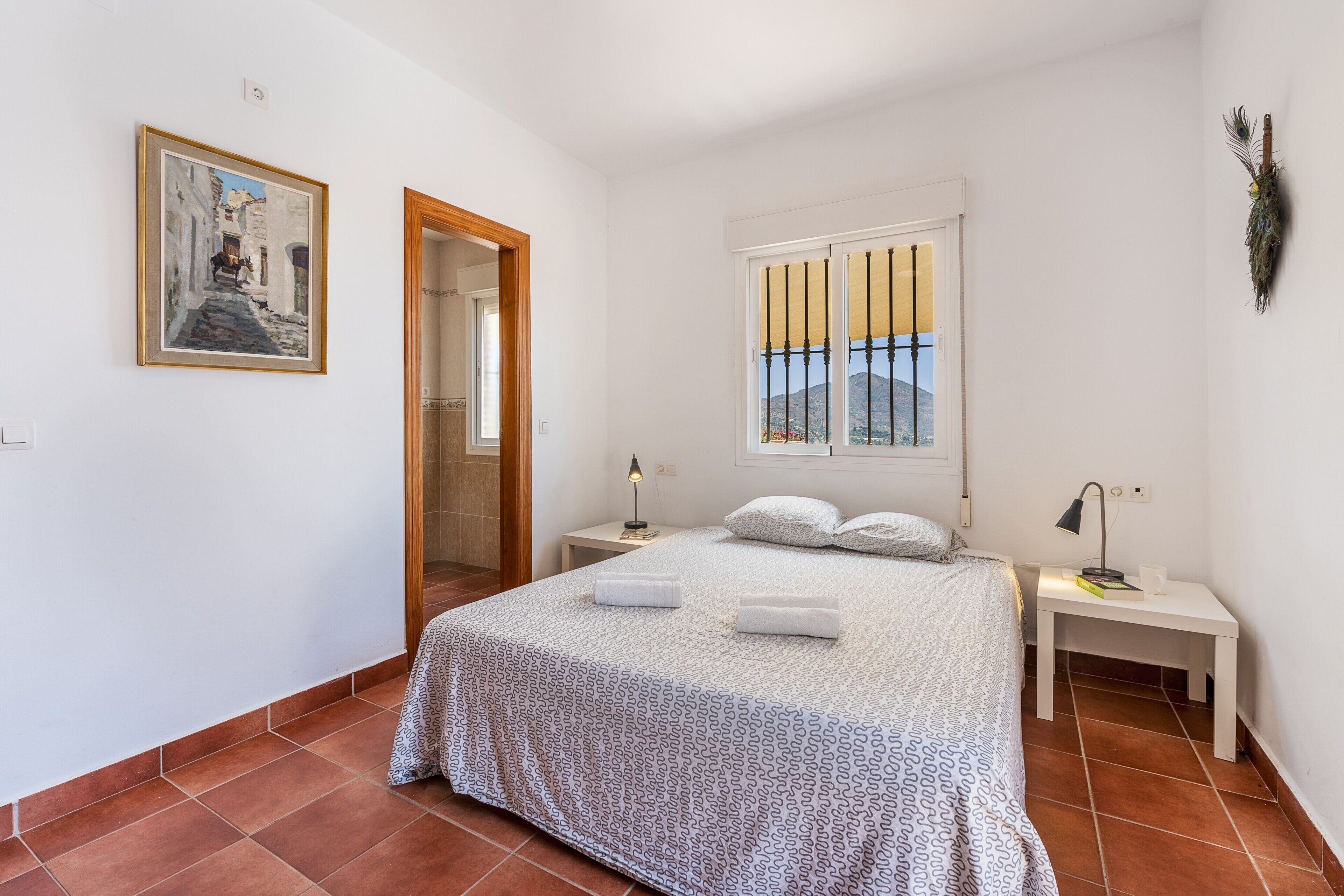 A bedroom with a bed, a nightstand, a bedside table, a window with a view, and a framed painting on the wall.
