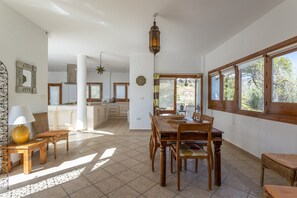 Un'ampia sala da pranzo con tavolo e sedie in legno, una cucina sullo sfondo e ampie finestre con vista sul verde.