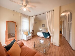 A hotel room with a bed, a sitting area with a sofa, a glass coffee table, a ceiling fan, and a wardrobe.