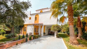 A two-story building with a white exterior and yellow accents, surrounded by a well-maintained garden with palm trees and various shrubs. - Nice Villa in Nueva Andalucia with Mountain View, Pool, Wi-Fi, A/C, Terraces & Garden (Nueva Andalucía)