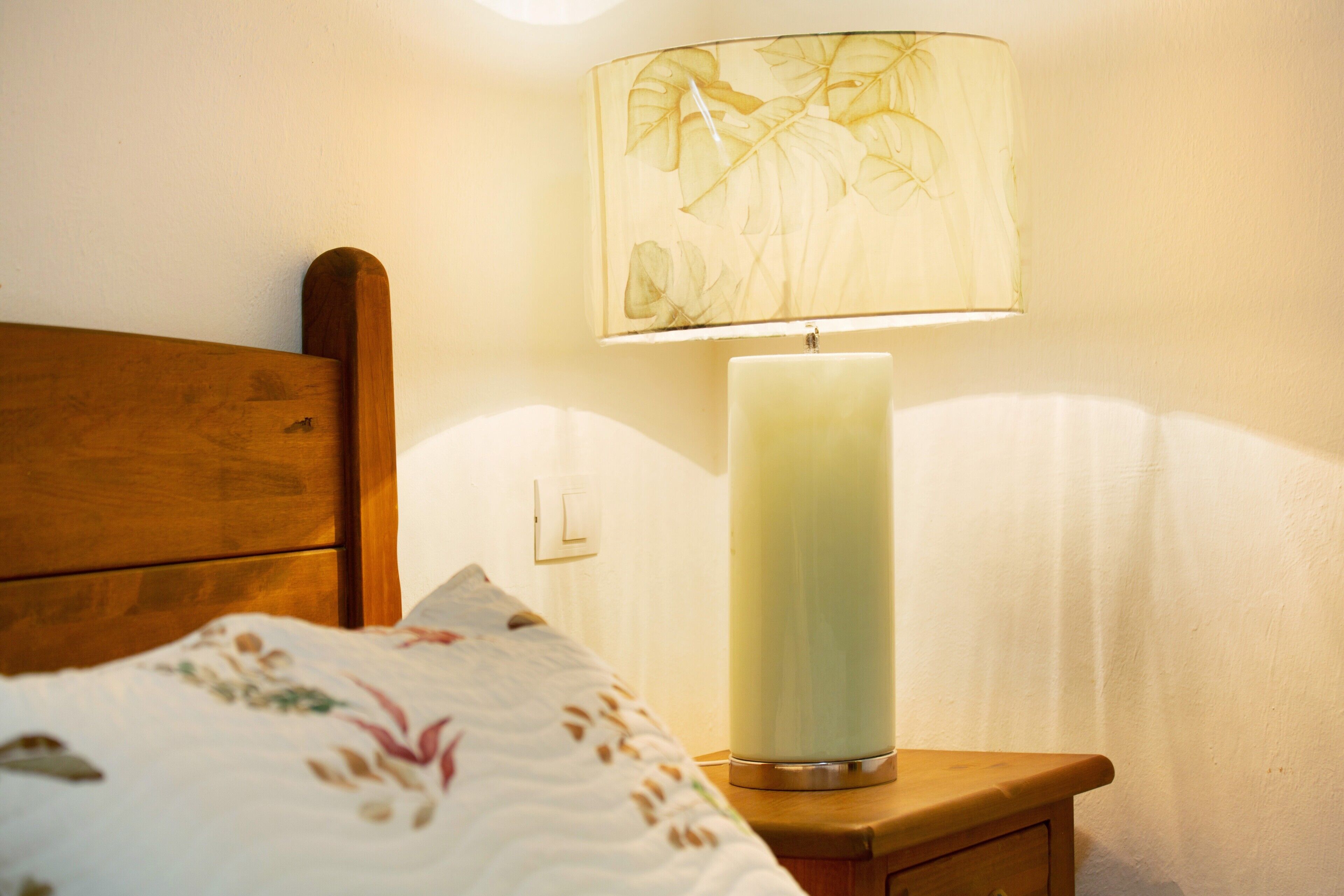 Ein Bett mit floralem Überwurf, ein Holz-Kopfstück, ein Nachttisch mit einer weißen Kerze und eine Wandlampe mit Blattformdesign.