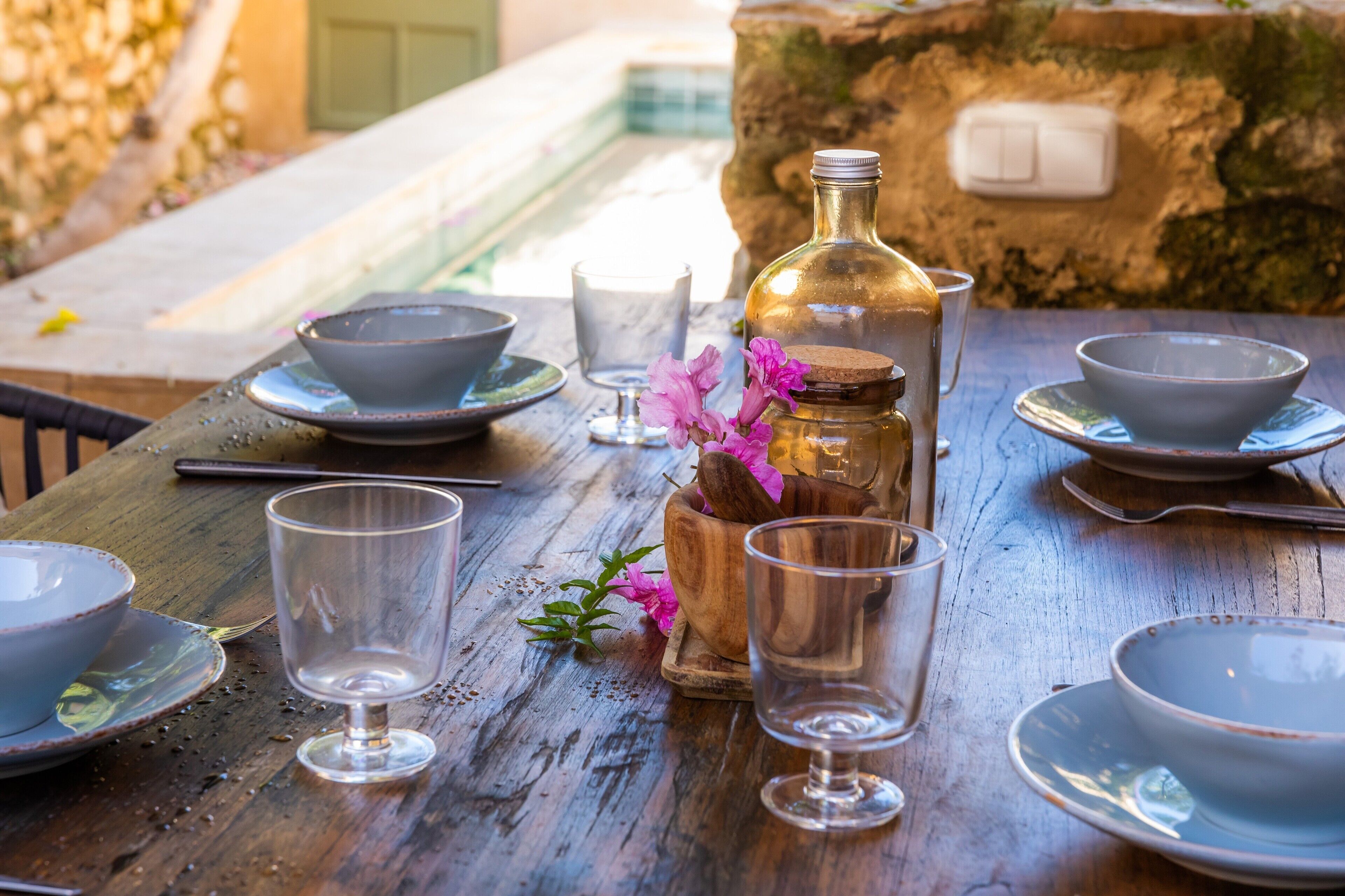 Une table en bois, sur laquelle sont disposés des bols, des verres et une bouteille, est placée devant un mur de pierre avec un interrupteur.