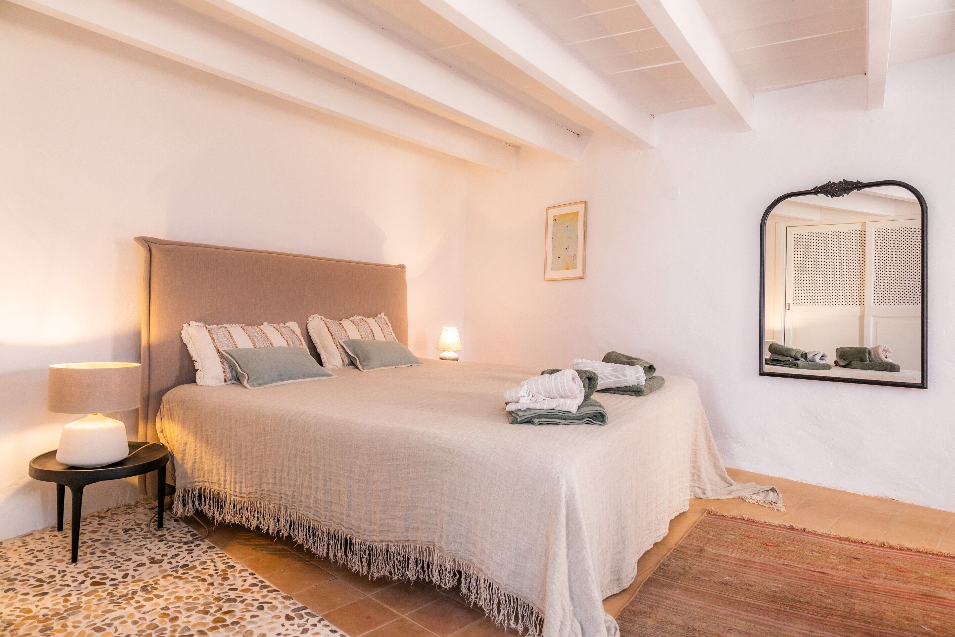 Een slaapkamer met een bed, nachtkastje, lamp, spiegel en vloerkleed.
