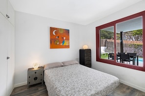Une chambre à coucher comprenant un lit, une table de chevet, une commode, un tableau et une fenêtre donnant sur un espace extérieur.