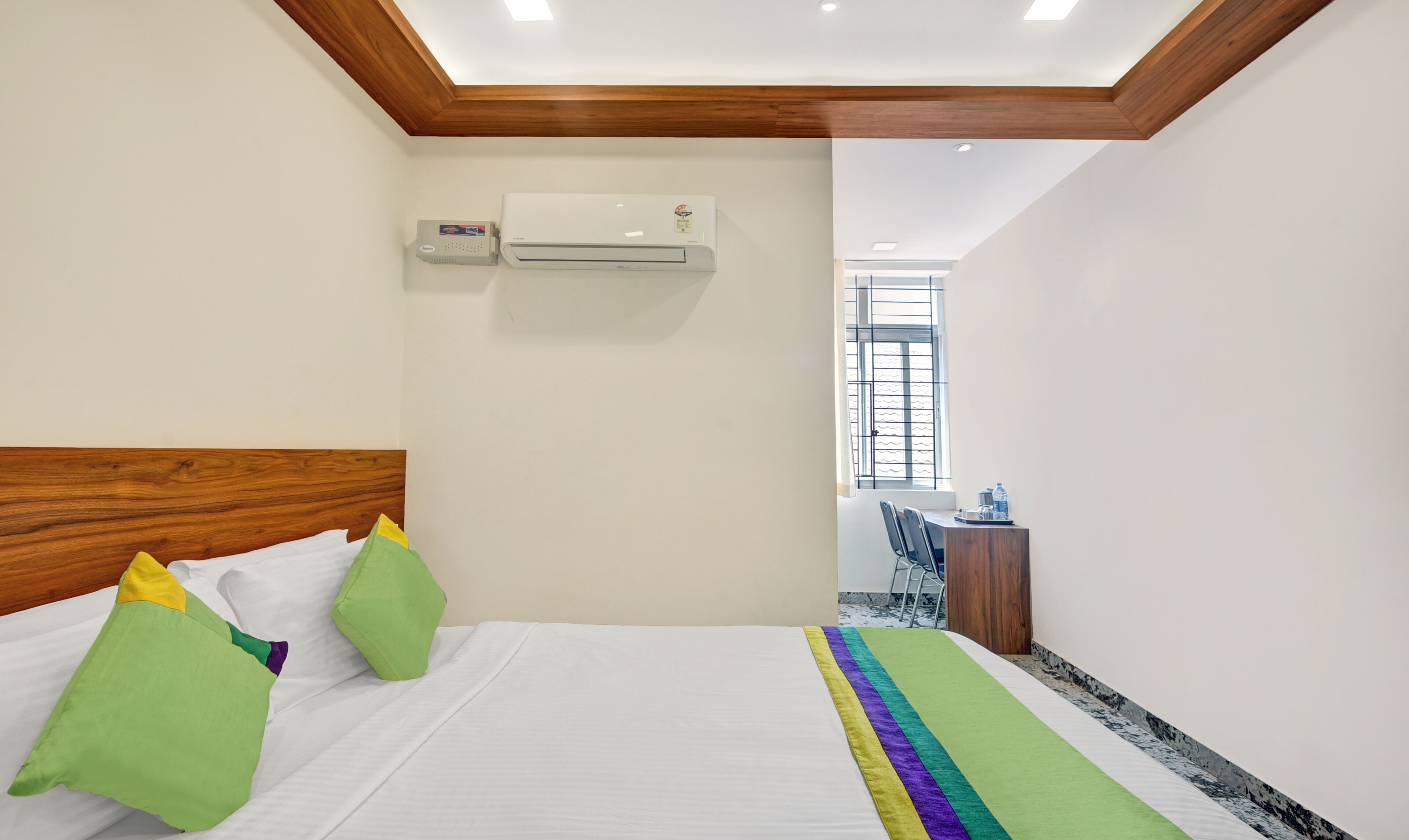 A hotel room with a wooden headboard, a bed with white bedding and colorful pillows, an air conditioning unit, a small desk with a chair, and a window with bars.