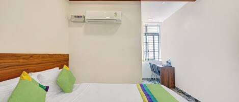 A hotel room with a wooden headboard, a bed with white bedding and colorful pillows, an air conditioning unit, a small desk with a chair, and a window with bars.