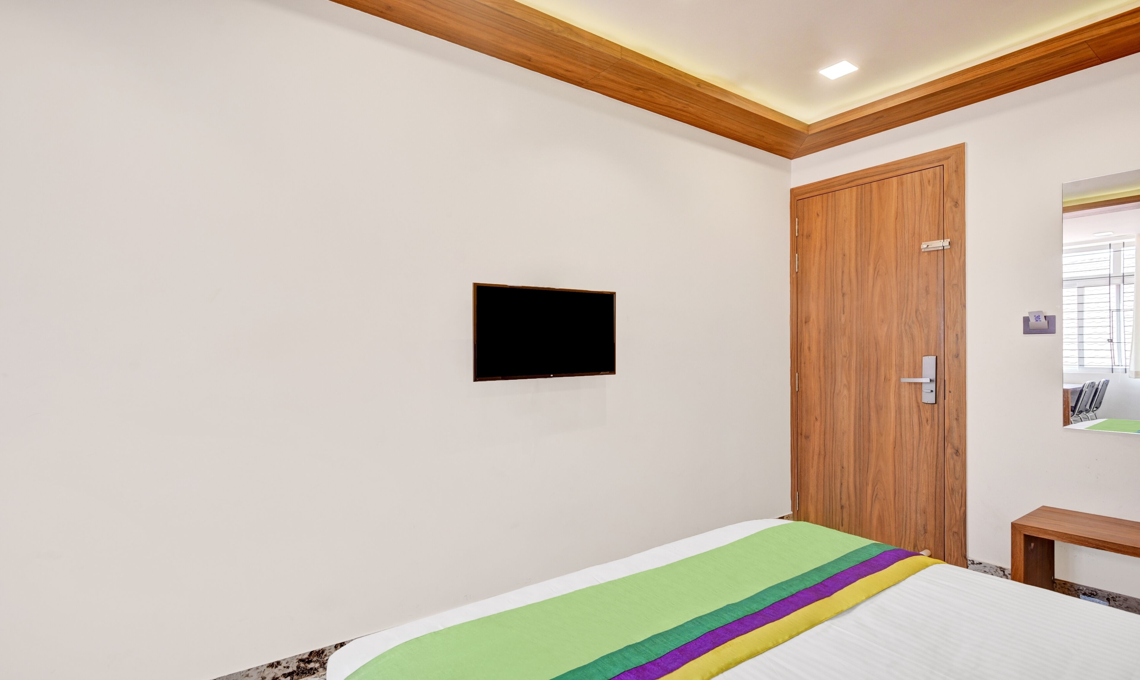 A bedroom with a bed, a wooden door, a mounted TV, and a small bedside table.