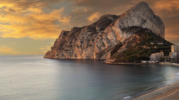 Un paisaje costero con una imponente montaña rocosa, mar tranquilo y algunos edificios en la orilla.