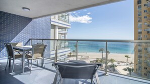 Ein Balkon mit Blick auf Strand und Meer, ein Esstisch mit Stühlen und ein modernes Gebäude im Hintergrund.