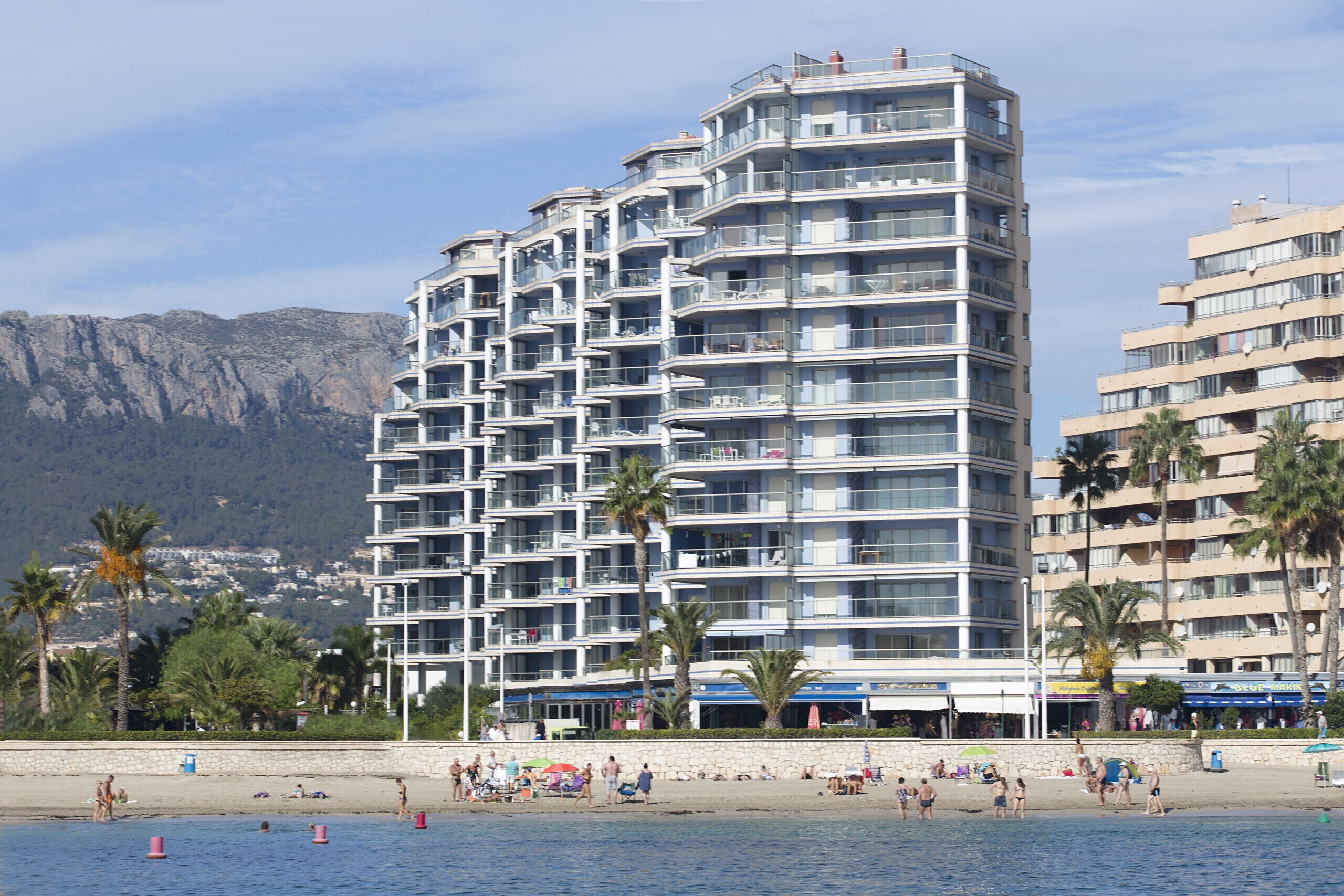 A multi-story hotel with balconies, located near a beach with palm trees and a mountainous backdrop.