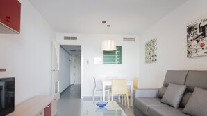Een moderne woonkamer met een grijze bank, een glazen salontafel en een flatscreen-tv op een houten kast. Er is een eethoek met een witte tafel en stoelen, en een keuken met witte kasten en een rode accentkleur.