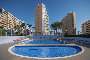 A swimming pool surrounded by tall residential buildings, palm trees, and a clear blue sky.