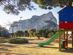 Un parque infantil con un tobogán, rodeado de árboles y una montaña al fondo.
