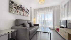 A modern living room with a grey sofa, a glass coffee table, a TV on a wooden cabinet, and a large window with sheer curtains. - First line / Wifi / Spa / Seaviews / Heated pool / Pets (Calpe)