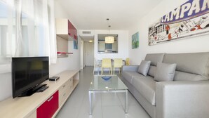 Una sala de estar moderna con un televisor de pantalla plana, una mesa de centro de vidrio, un sofá gris y un comedor al fondo.