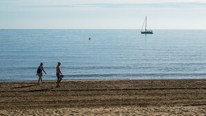 A beach with two people walking, a sailboat in the distance, and a clear sky.