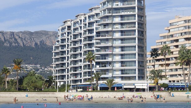 A multi-story hotel with balconies, located near a beach with palm trees and a mountainous backdrop.