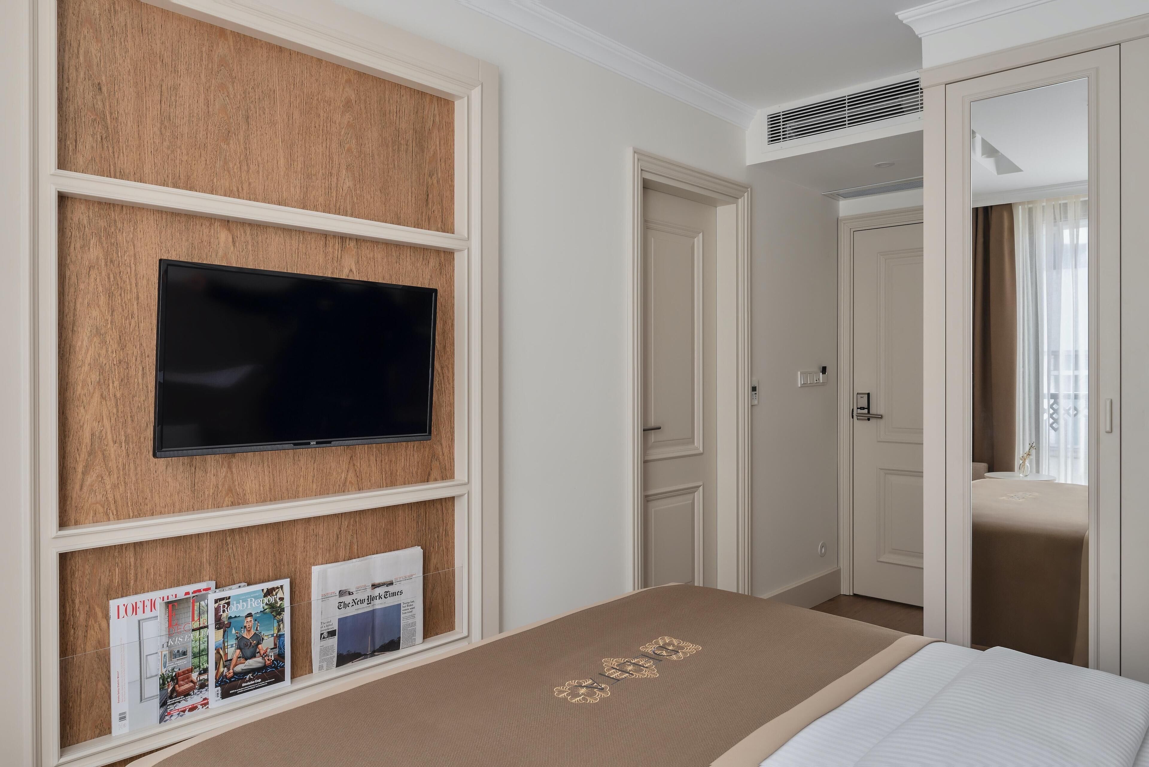 A hotel room with a bed, a mounted TV, and magazines on a shelf.